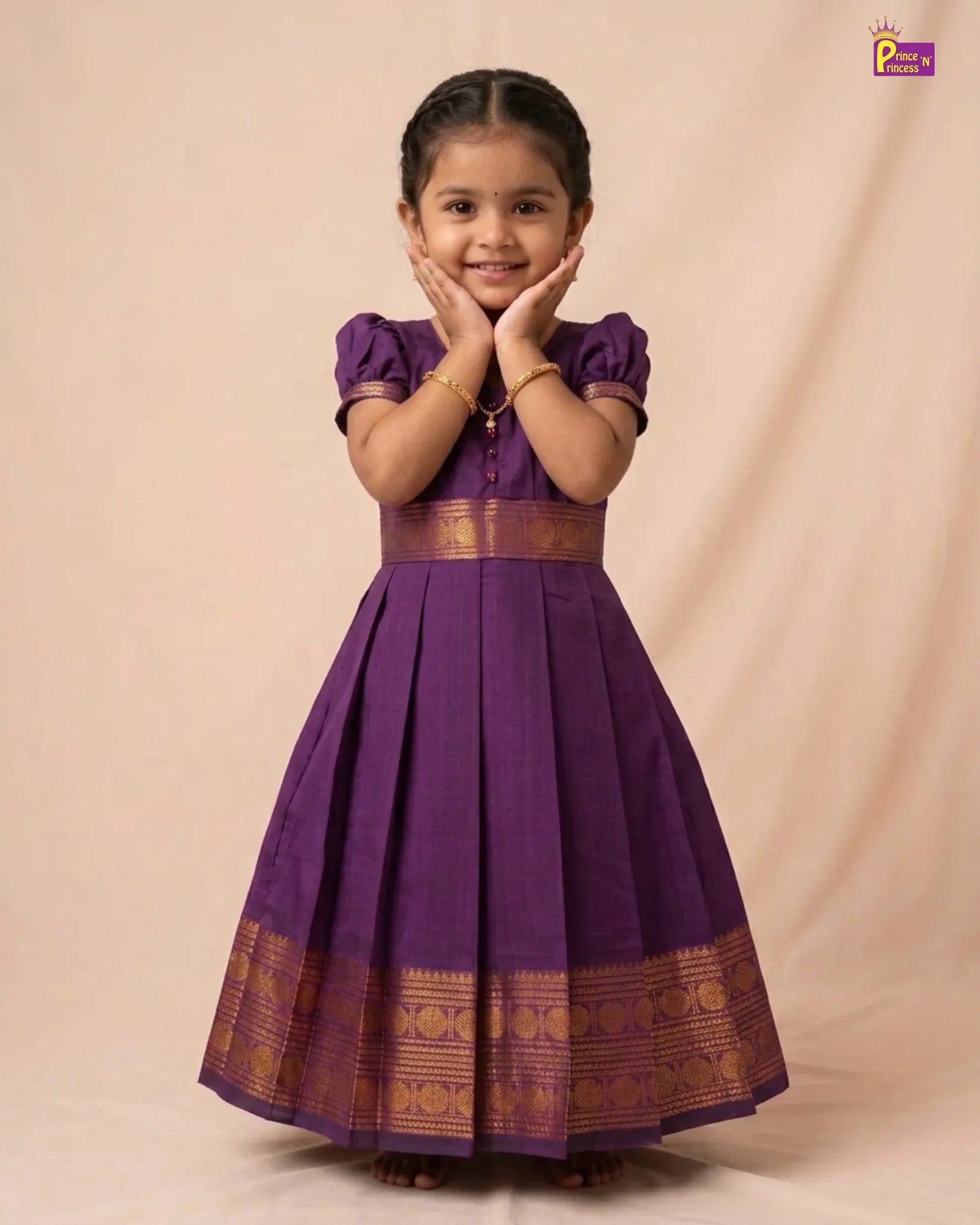 A young girl modeling a purple traditional Indian dress with gold zari borders, standing against a beige studio backdrop.