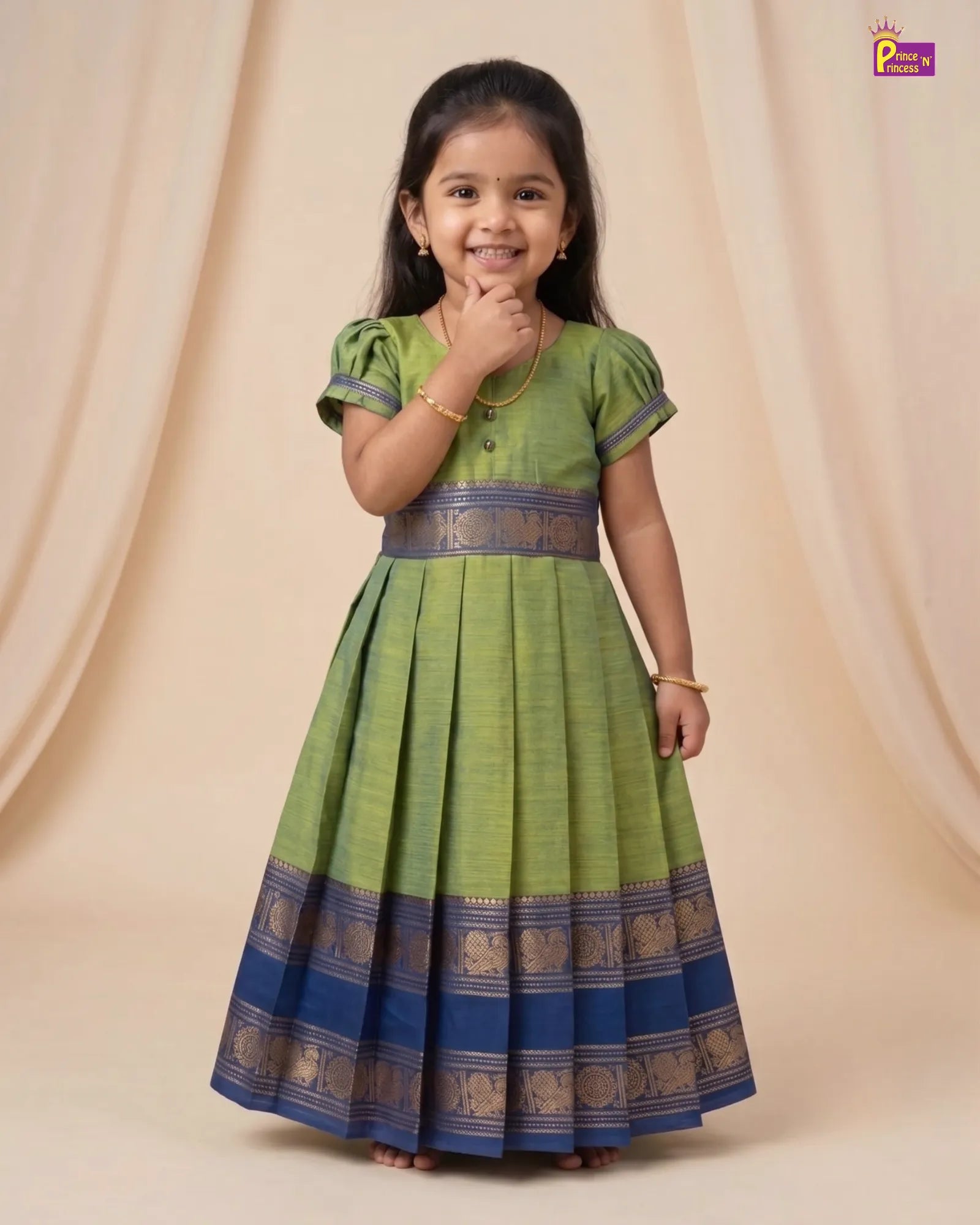 A young girl modeling a green and blue traditional Indian dress with gold zari borders, standing against a beige draped studio backdrop.