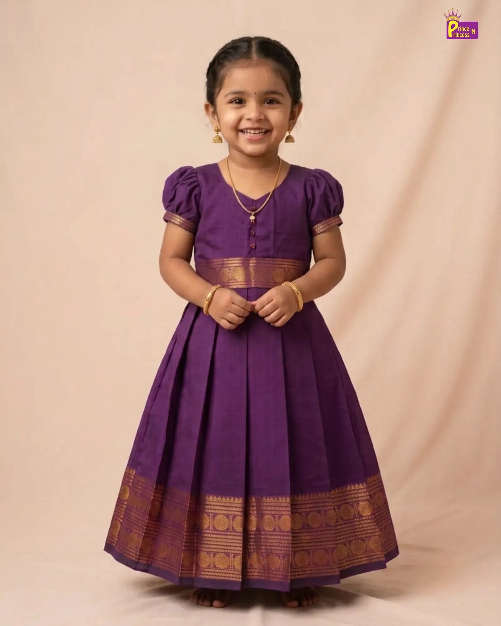 A young girl wearing a purple traditional dress with gold zari borders, standing against a beige studio backdrop.