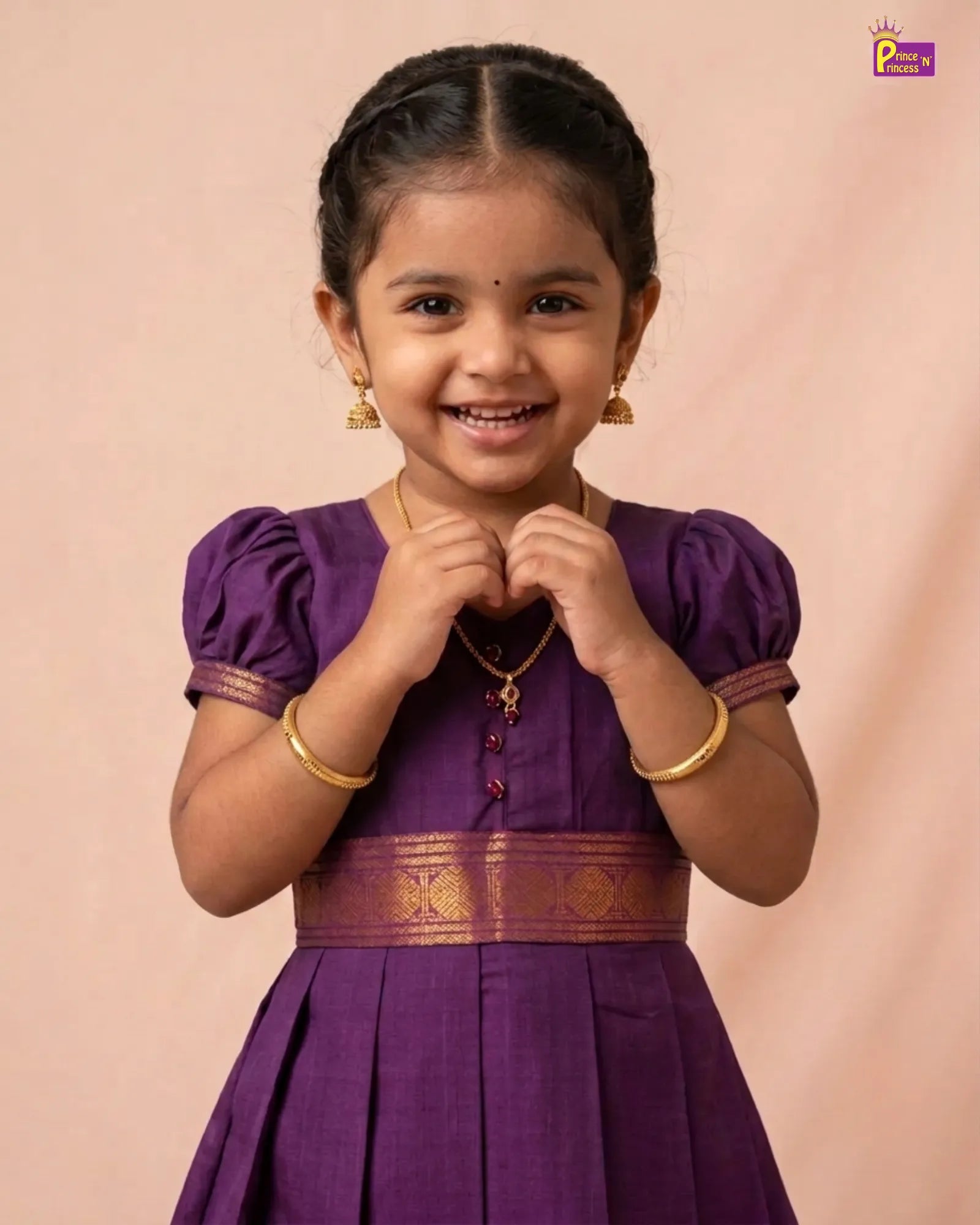 A young girl smiling and posing in a traditional purple dress with gold jewelry, set against a solid light peach background.