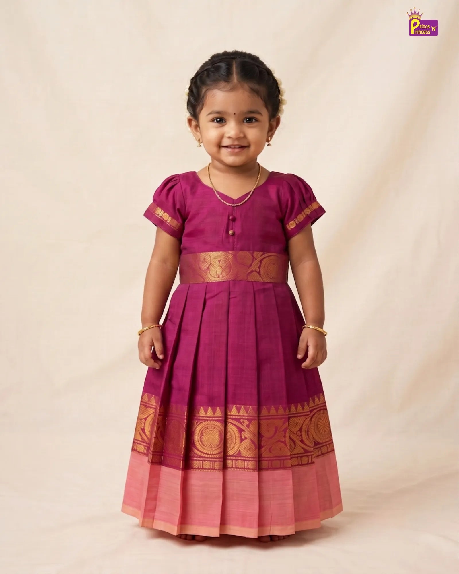 A young girl modeling a magenta pink traditional Indian salwar suit with gold zari embroidery, standing against a plain beige backdrop.