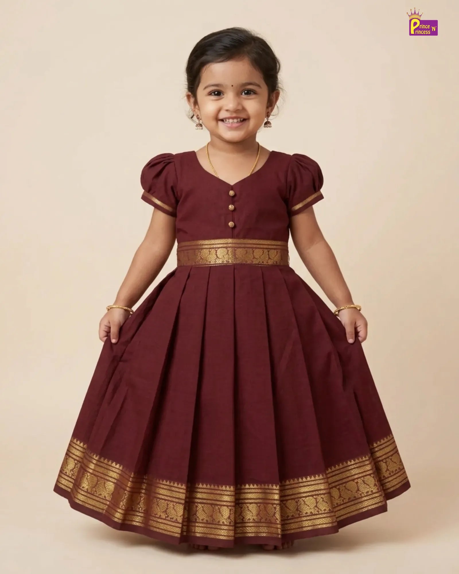 A young girl wearing a maroon A-line party dress with gold zari borders and button details, standing against a solid beige background.