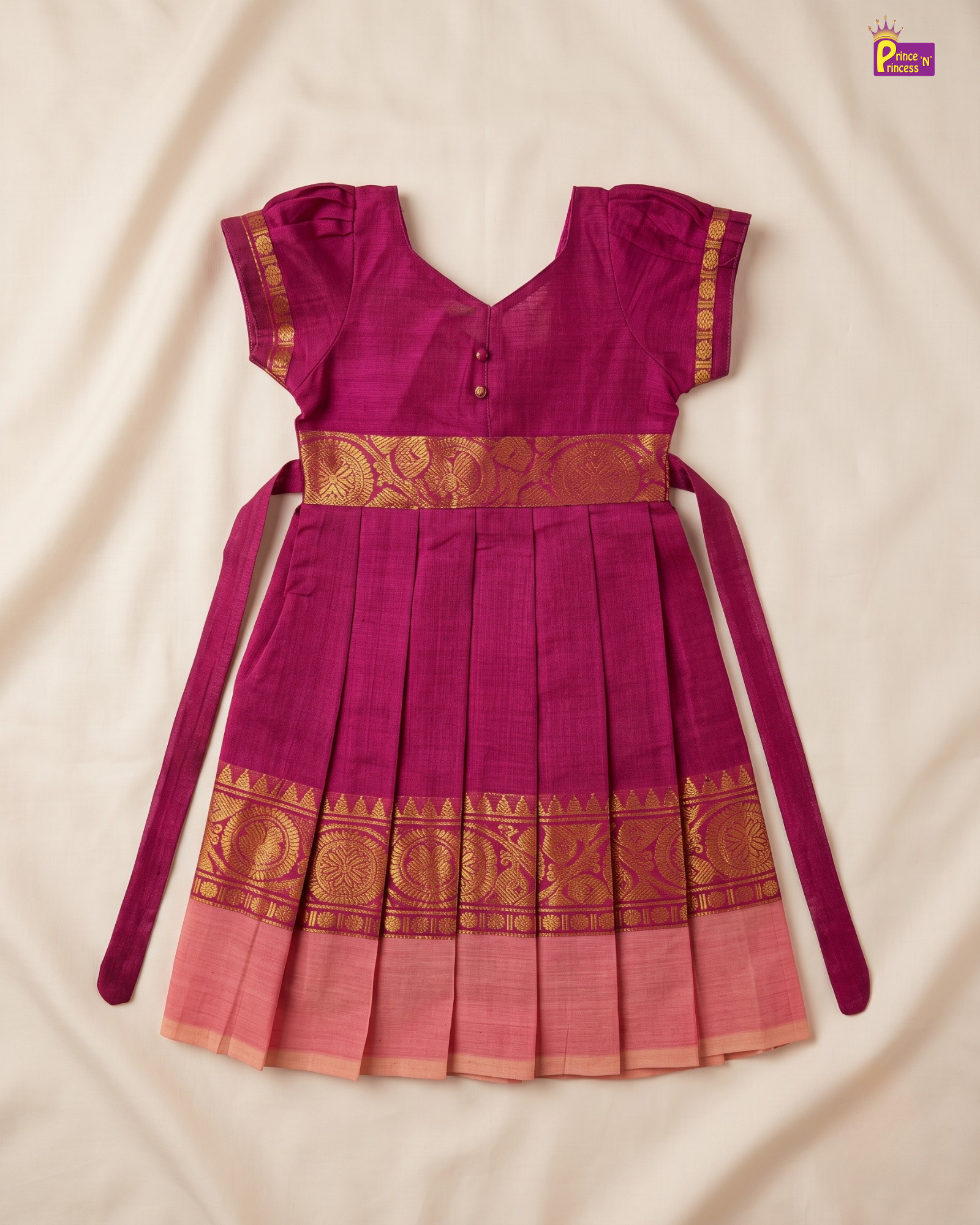 A magenta pink traditional Indian dress with gold zari embroidery, featuring a pleated skirt, short sleeves, and a waist tie, laid flat on a cream-colored fabric background.