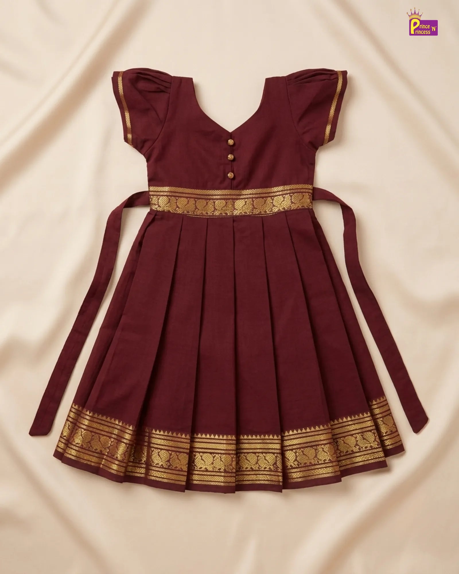 A maroon pleated baby dress with gold patterned borders and a matching tie belt, laid flat on a cream-colored fabric background.