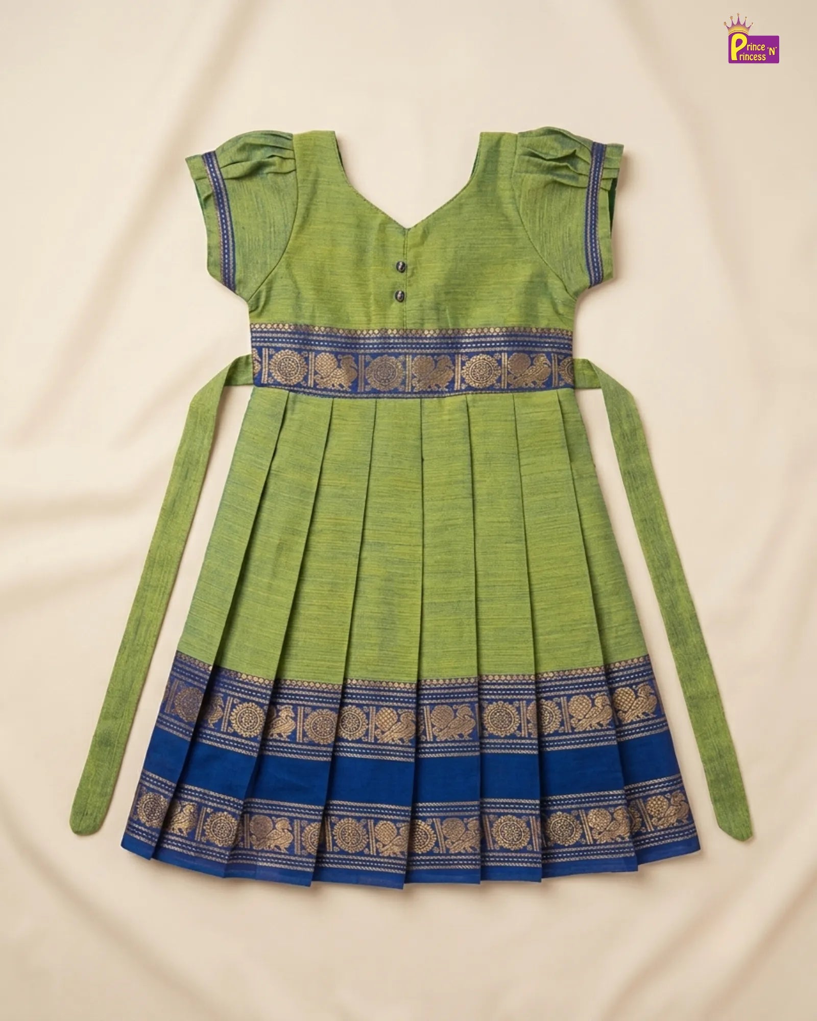 A green and blue traditional Indian girl's dress with pleated skirt, short ruffled sleeves, and intricate gold zari borders, laid flat on a cream fabric background.