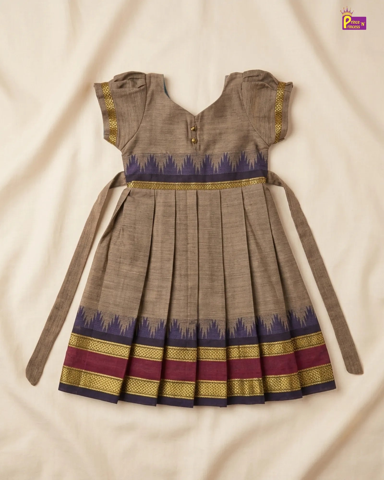A traditional Indian girl's dress in brown fabric with a blue and gold patterned waistband and hem, laid flat on a cream-colored fabric background.