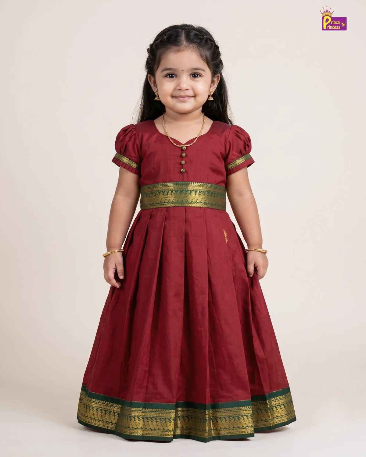 A young girl modeling a traditional Indian maroon red Anarkali gown with gold and green zari borders, standing against a plain beige studio background.