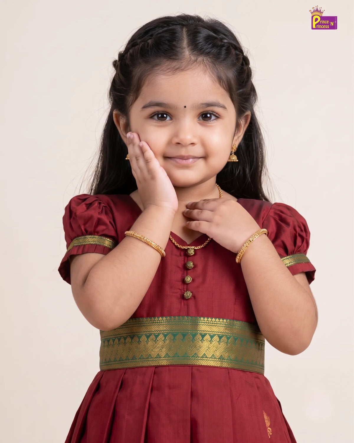 A young girl with dark hair in a braided style, wearing a traditional Indian maroon dress with gold and green borders, posing against a light beige background.