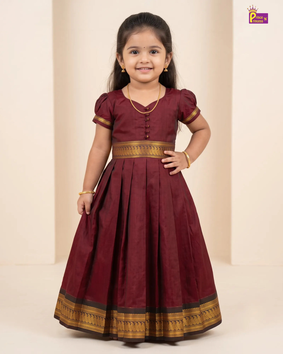 A young girl modeling a maroon Anarkali-style dress with gold and black zari borders, standing against a beige studio backdrop.