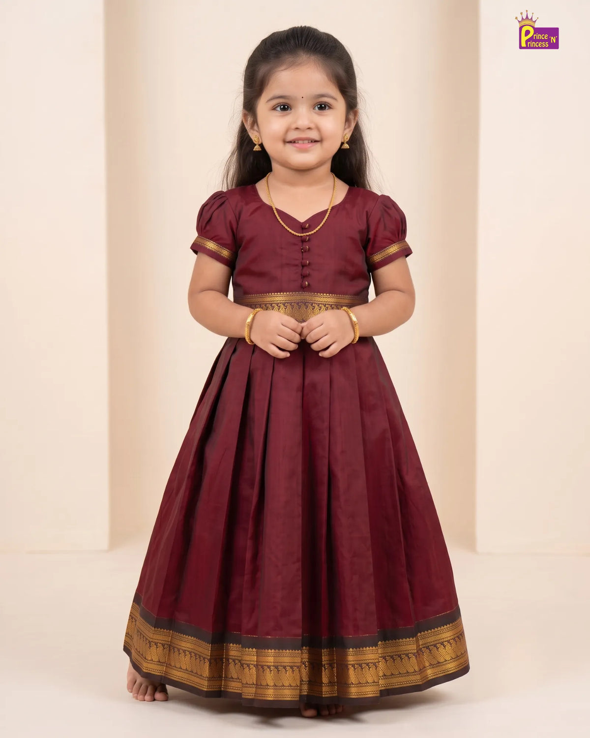 A young girl wearing a traditional Indian maroon Anarkali gown with gold and black border detailing, standing against a beige background.