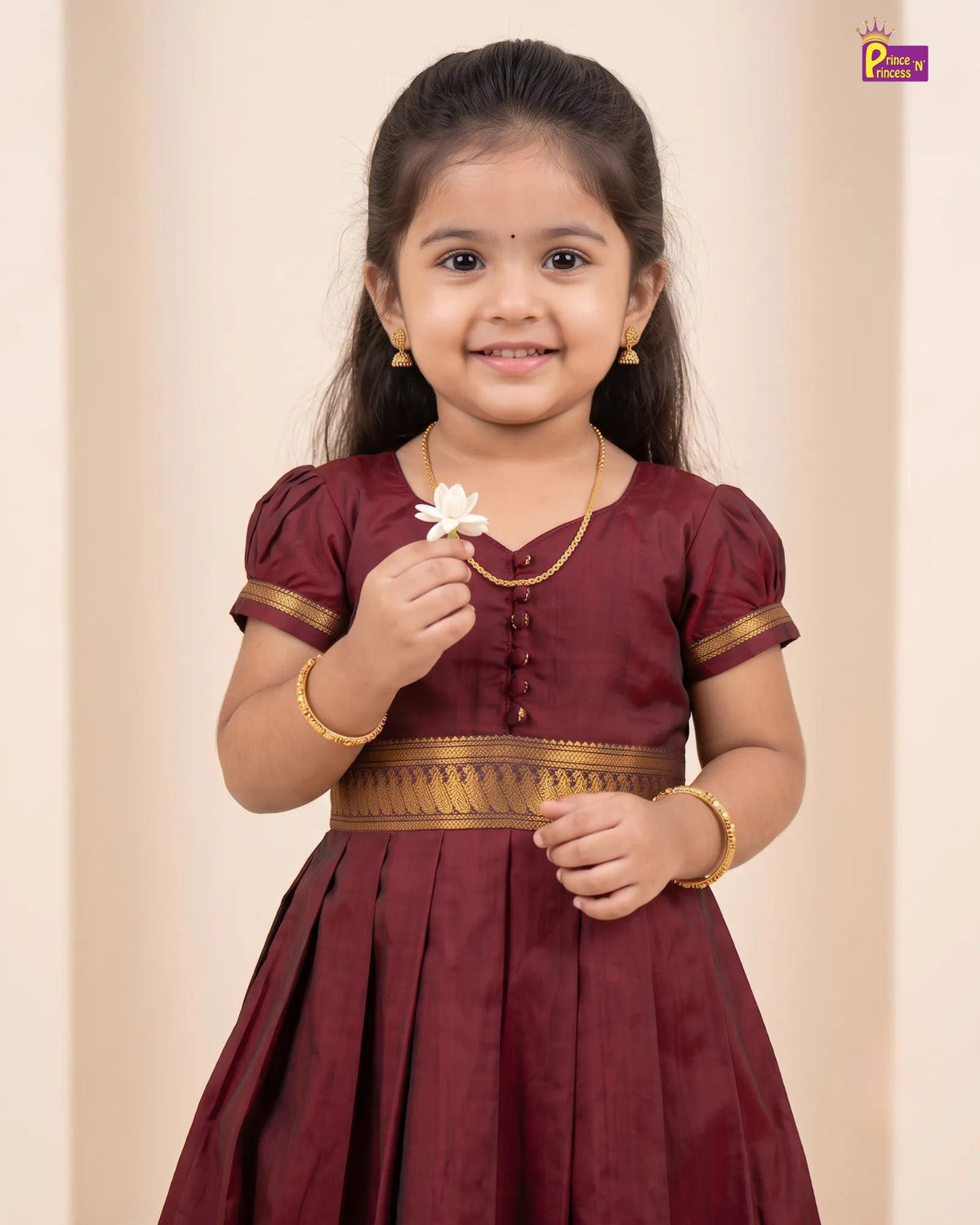 A young girl with dark hair wearing a maroon traditional dress with gold trim, holding a small white flower. The background is a soft beige studio backdrop.
