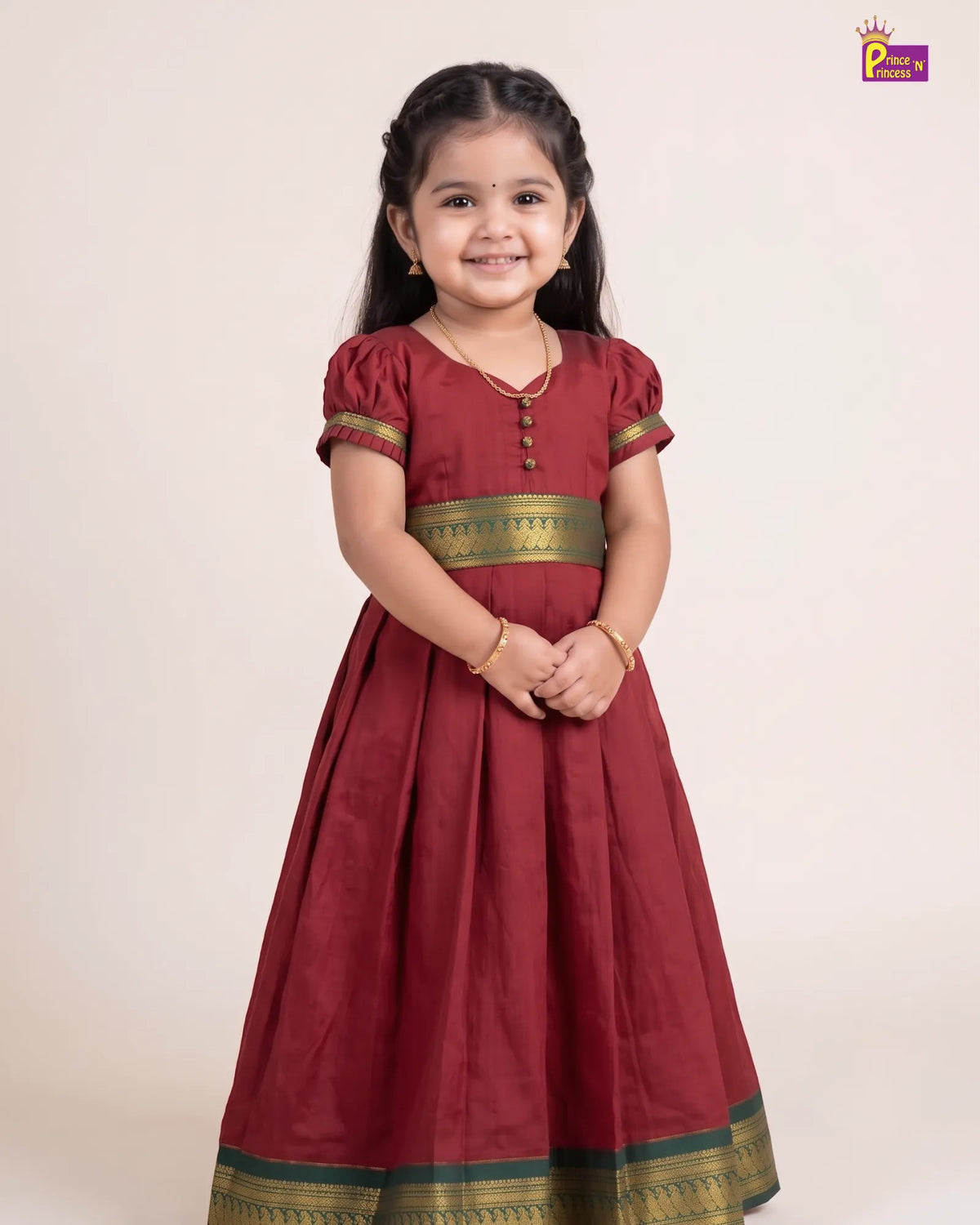A young girl with dark hair wearing a traditional maroon red Anarkali gown with gold and green borders, standing against a plain beige studio background.