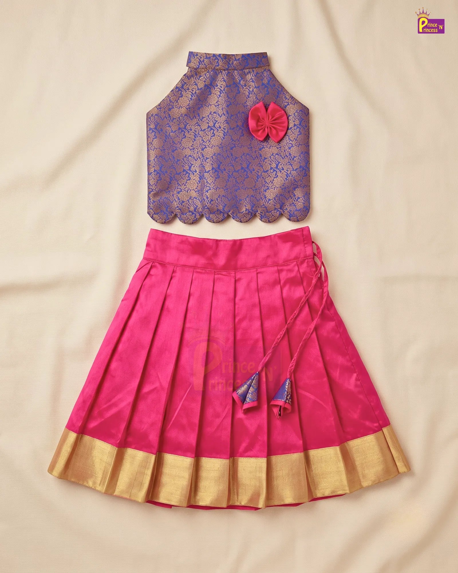 A traditional Indian children's two-piece outfit laid flat on a beige fabric background. The set includes a sleeveless purple lace blouse with a pink fabric flower accent and a matching pleated pink skirt with a gold zari border and tassel ties.