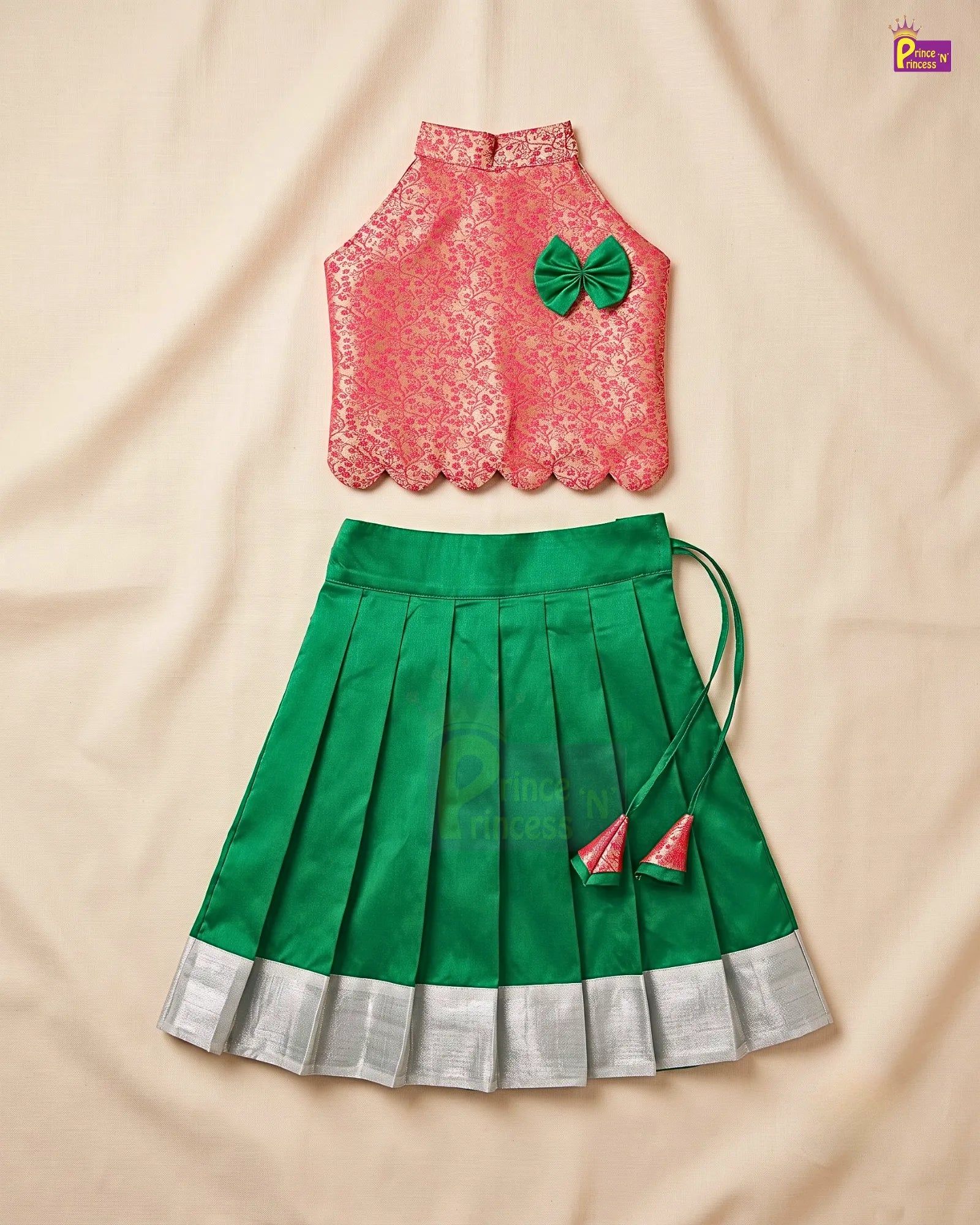 A two-piece girls' traditional outfit laid flat on a beige fabric background. The top is a sleeveless coral-pink lace blouse with scalloped edges and a green bow accent.