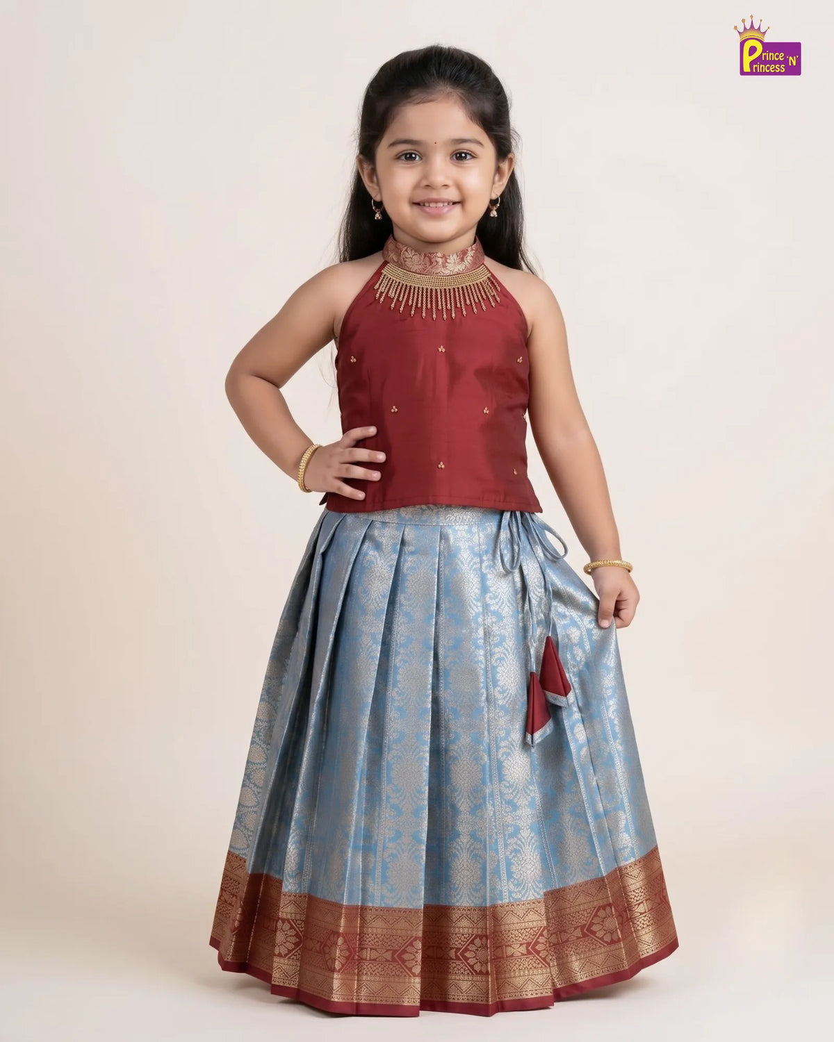 A young girl modeling a traditional Indian ethnic wear set consisting of a maroon sleeveless top with gold embroidery and a light blue pleated skirt with gold zari patterns and borders, against a plain beige background.