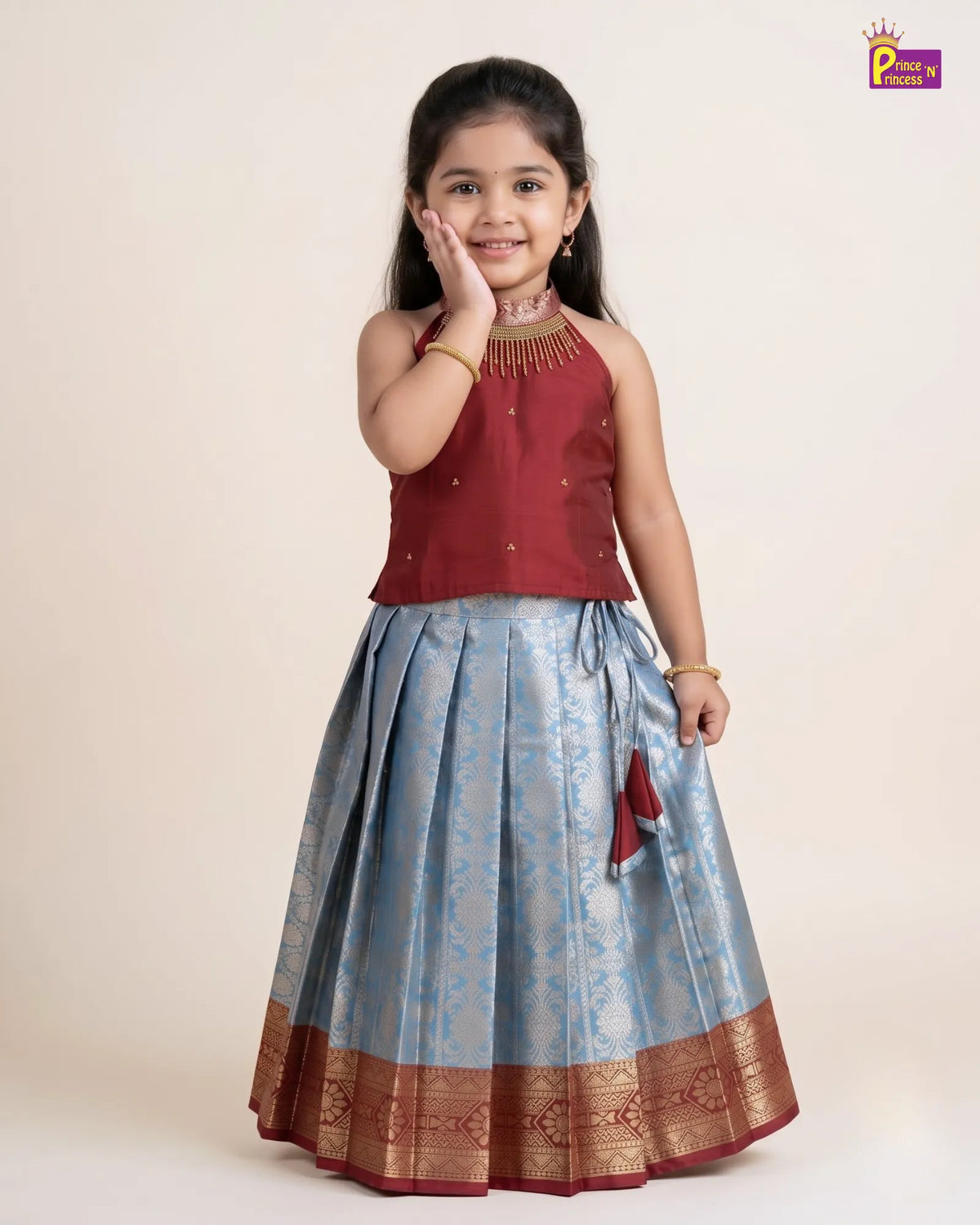 A young girl modeling a traditional Indian outfit consisting of a maroon sleeveless top with gold embroidery and a light blue pleated skirt with gold zari borders, set against a plain beige background.