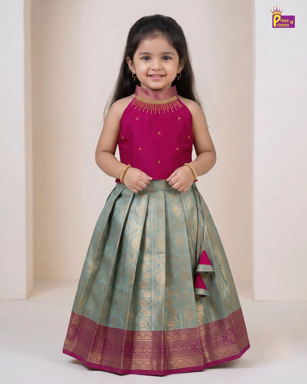A young girl modeling a traditional Indian lehenga choli dress. The sleeveless magenta pink choli has gold embroidery, and the pleated skirt is a sage green with gold zari motifs and a magenta border.