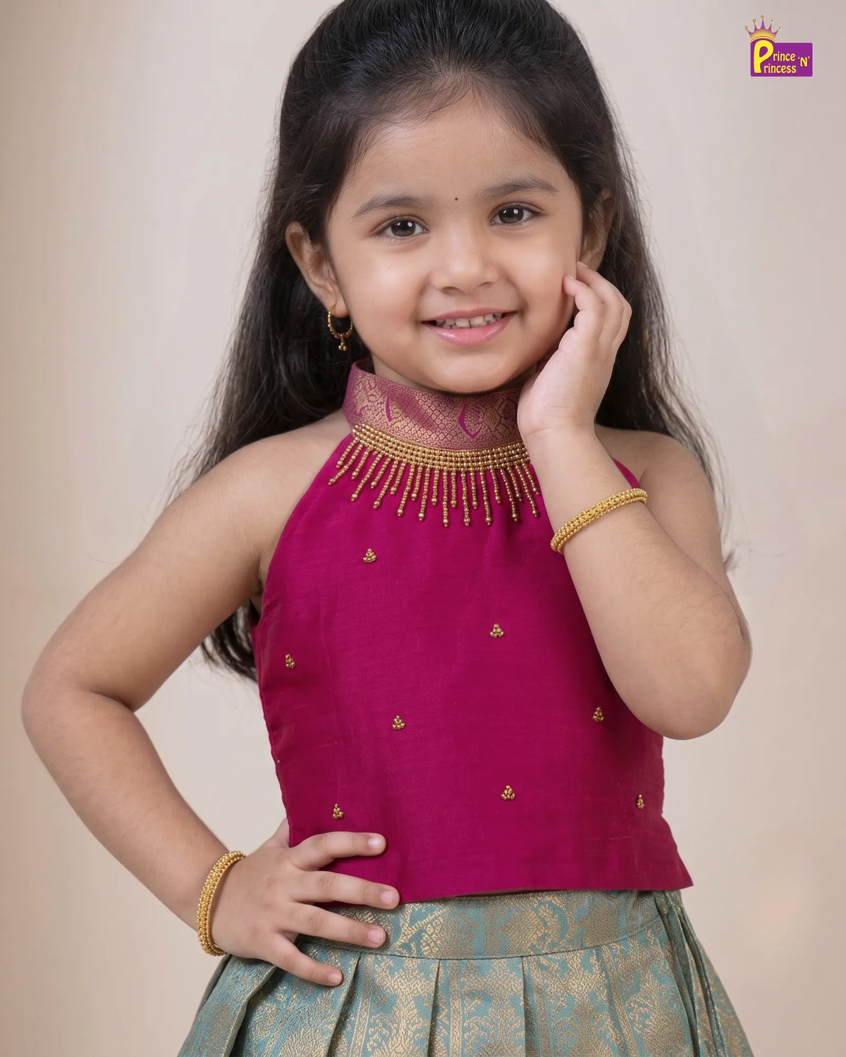 A young girl modeling a sleeveless magenta top with gold embroidery and a matching pleated skirt, set against a plain beige background.