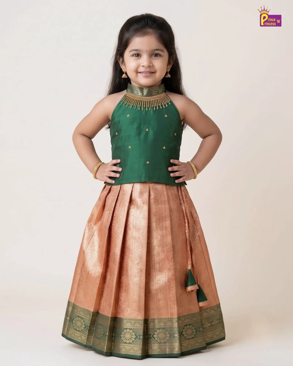 A young girl modeling a traditional Indian ethnic dress with a dark green sleeveless top and a pleated peach-beige skirt with gold zari embroidery and a green border, set against a plain light beige background.