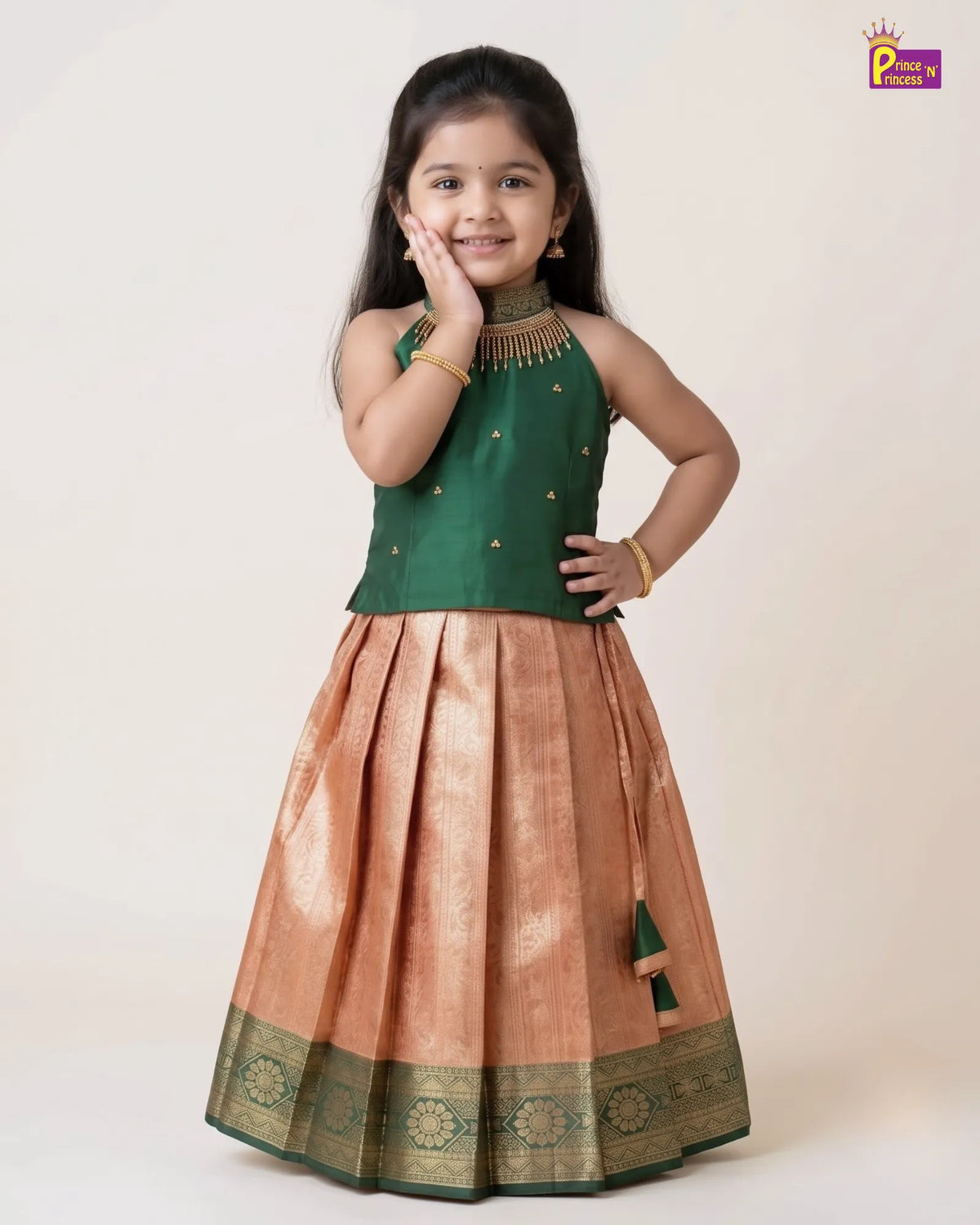 A young girl modeling a traditional Indian ethnic dress consisting of a dark green sleeveless top with gold embroidery and a pleated peach-beige skirt with a green and gold zari border, set against a plain light beige background.