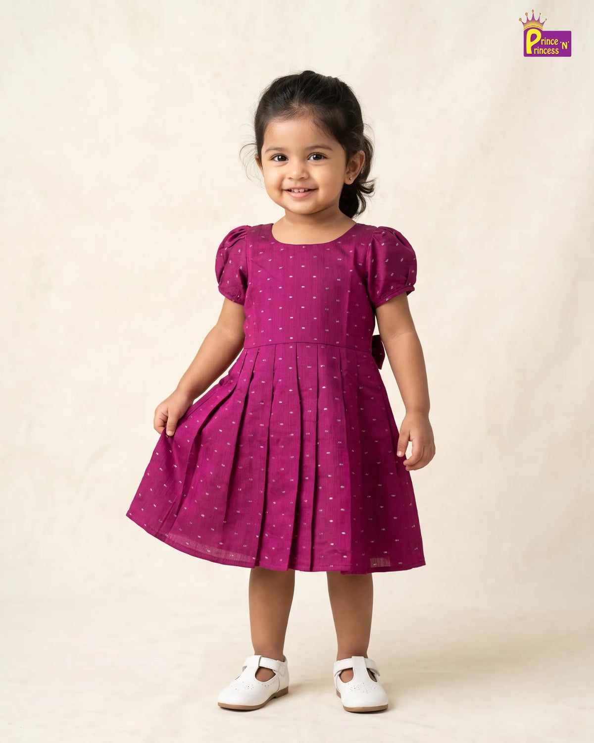 A young girl with dark hair tied back is wearing a magenta purple short-sleeve dress with a white polka dot pattern. She is standing against a warm beige textured backdrop, wearing white Mary Jane shoes.