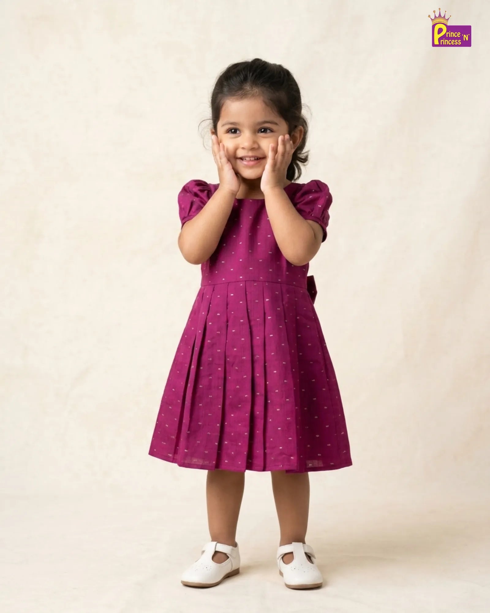 A young girl wearing a magenta purple short-sleeve dress with small white polka dots, standing against a warm beige textured backdrop. She is smiling and posing with her hands on her cheeks, wearing white Mary Jane shoes.
