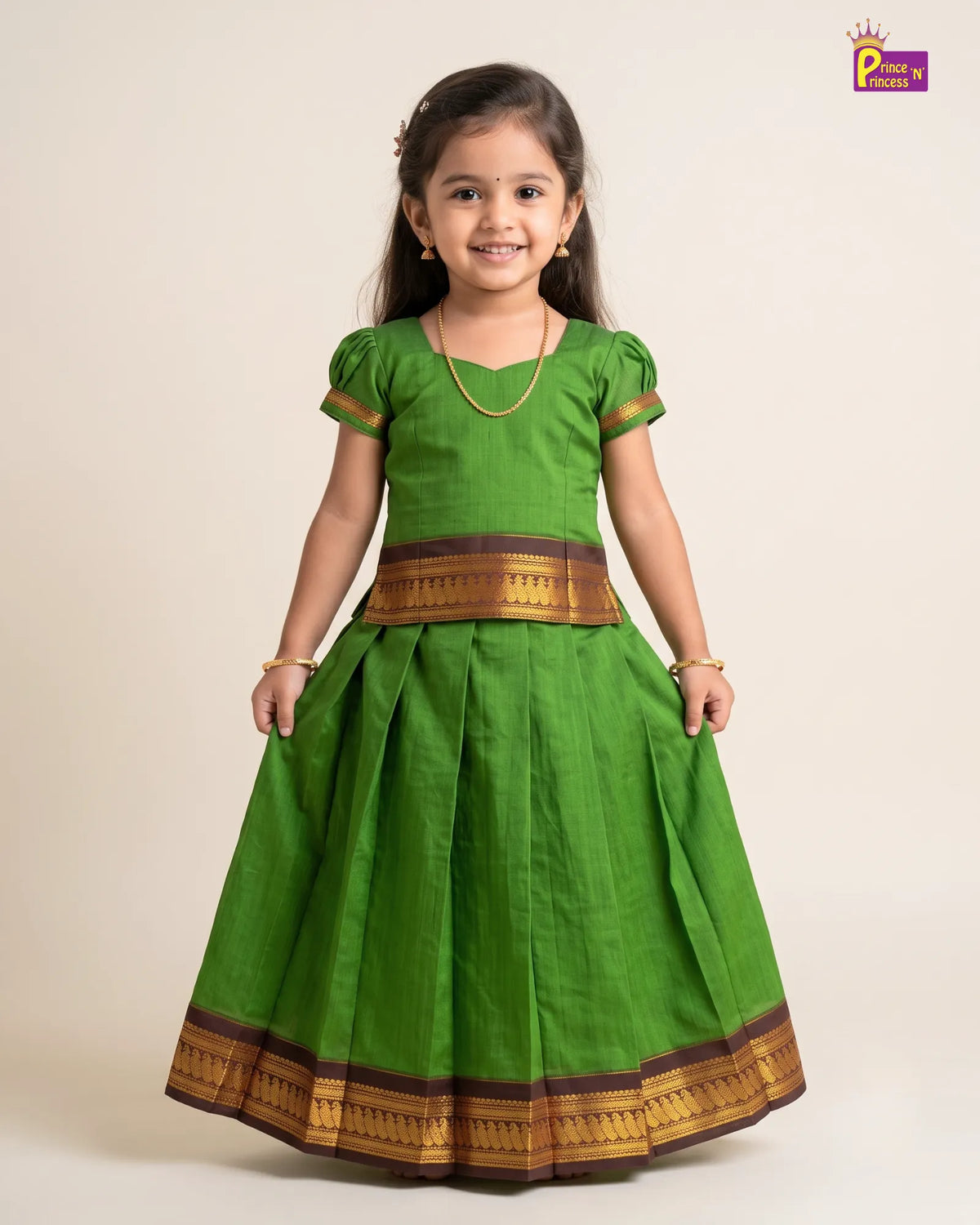 A young girl modeling a traditional green Indian dress with gold and brown borders, standing against a plain beige background.