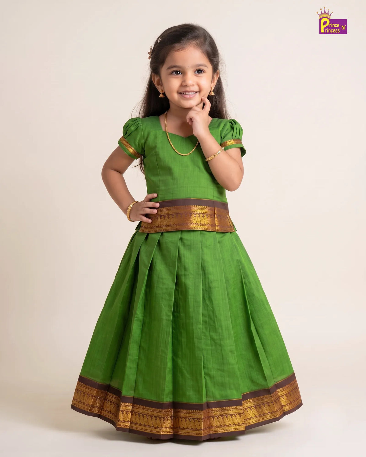 A young girl modeling a green traditional Indian dress with gold and brown zari borders, standing against a plain beige studio background.