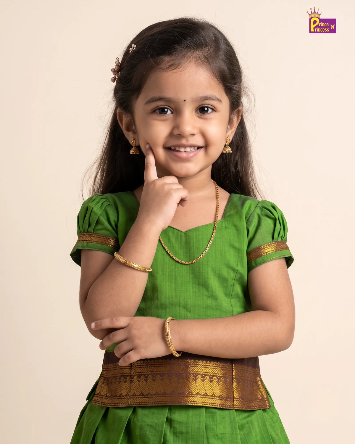 A young girl with dark hair wearing a traditional green Indian dress with gold trim, a gold necklace, and gold bangles, posing against a light beige background.