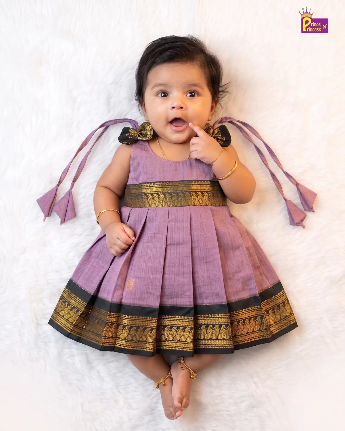 A baby girl lying on a white faux fur surface wearing a lavender purple traditional Indian dress with gold and black borders and matching ribbon ties. The brand logo 'Pancha's Creations' is visible in the top right corner.