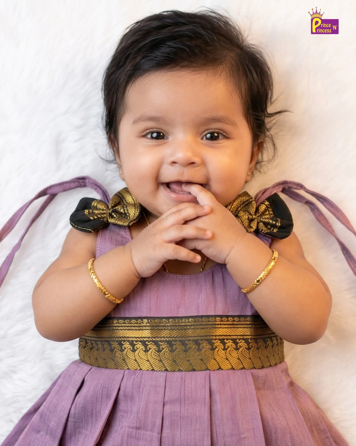 A baby girl wearing a lavender purple traditional dress with gold patterns and black bow details, lying on a white textured blanket.
