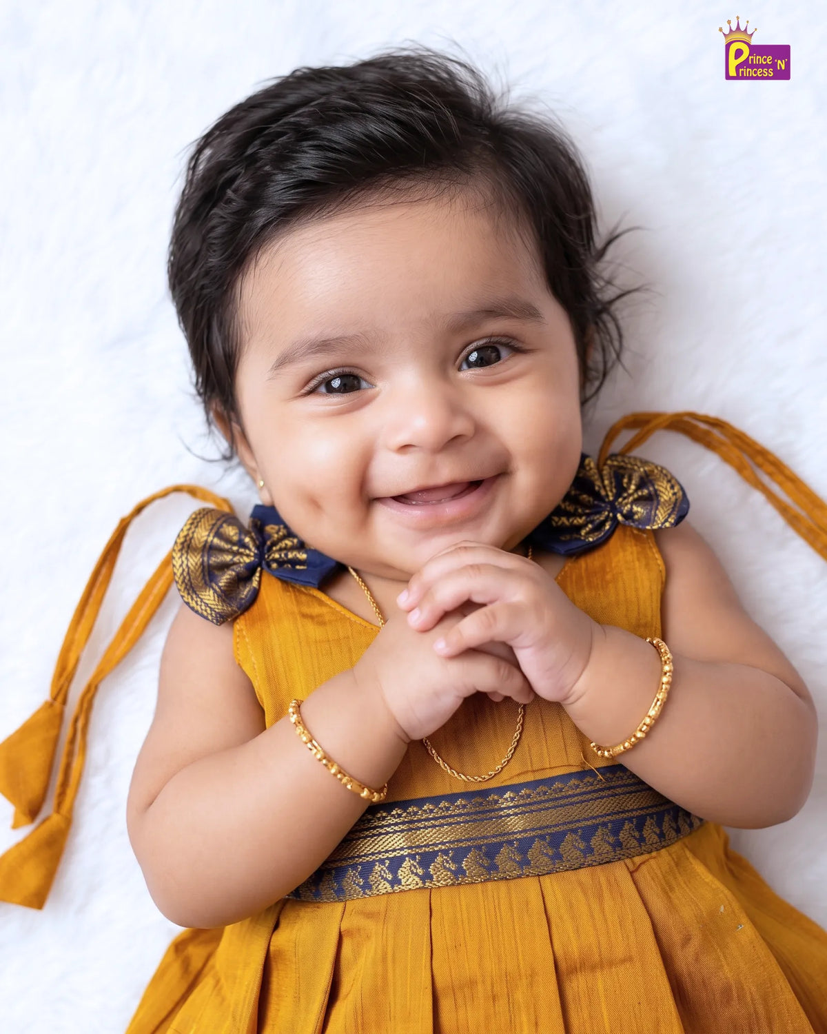 A baby girl lying on a white textured blanket, wearing a mustard yellow traditional dress with navy blue and gold borders, bow details on the shoulders, and matching ribbon ties. She is wearing gold jewelry including a necklace, bracelets, and ankle chains.