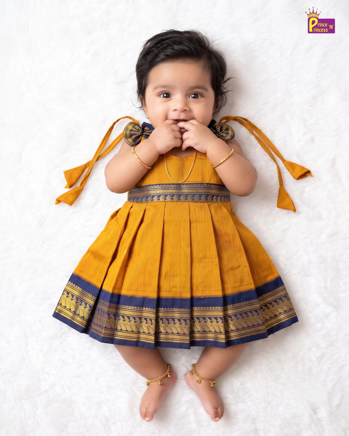 A toddler girl lying on a white textured blanket, wearing a mustard yellow pleated dress with navy blue and gold borders, gold bangles, and gold ankle bracelets.