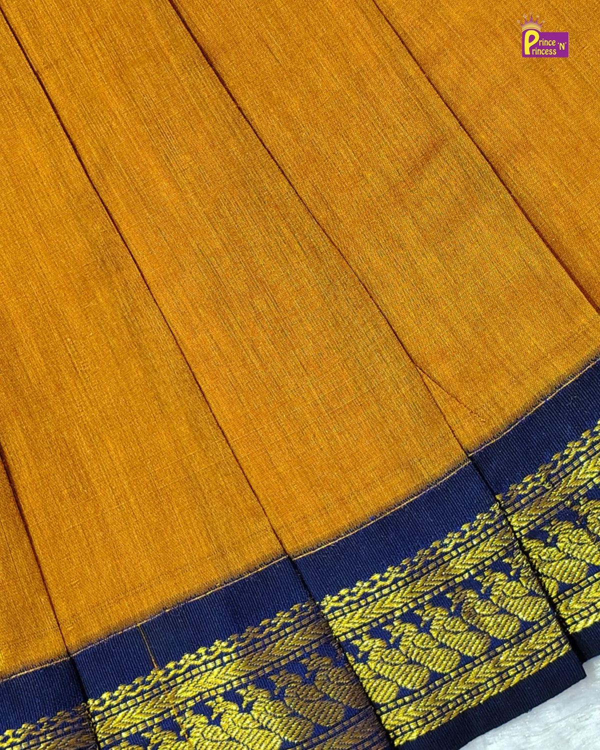 A smiling baby girl lying on a white textured blanket, wearing a mustard yellow dress with a navy blue and gold patterned waistband and bow details on the shoulders.