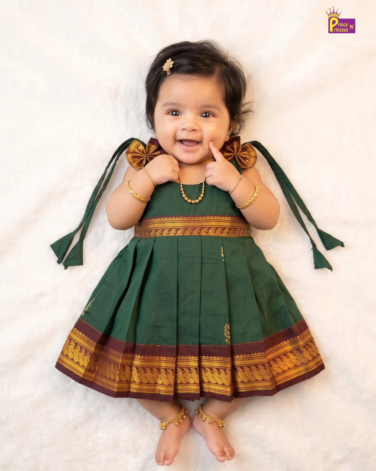 A baby girl wearing a dark green traditional dress with gold and maroon borders, lying on a white textured blanket. She is wearing gold jewelry and has a small gold hair clip.