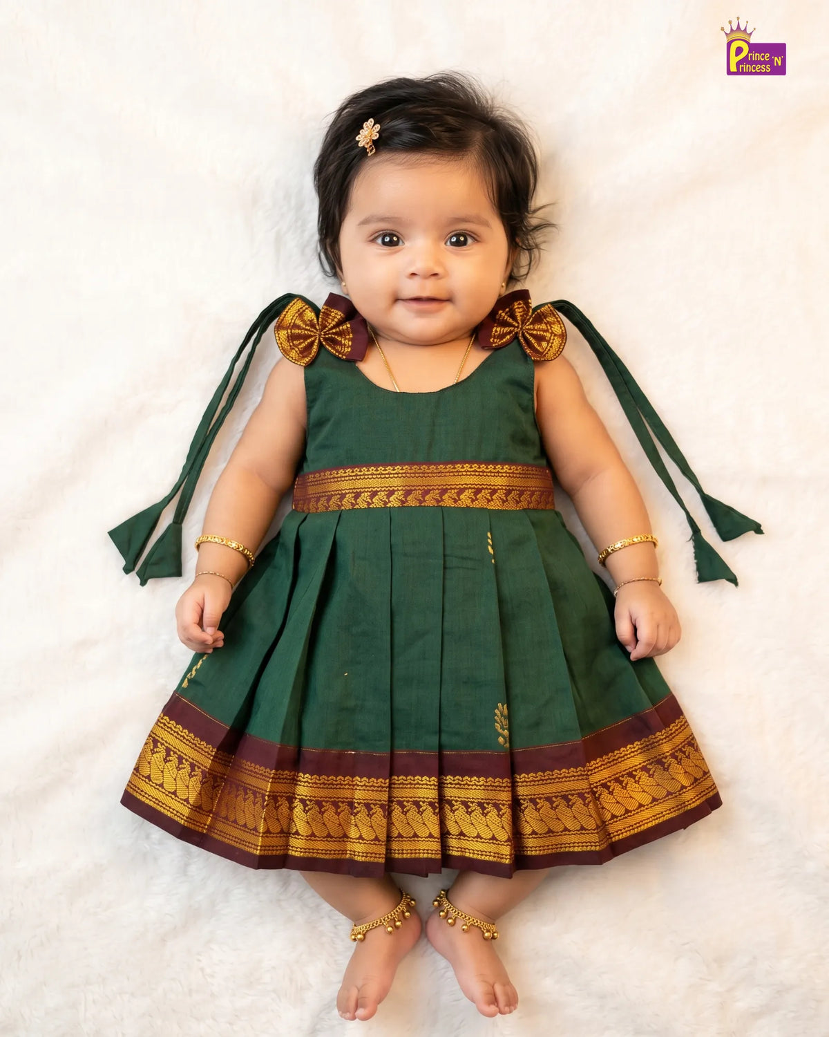 A baby girl wearing a dark green traditional dress with gold and maroon borders, lying on a white textured blanket. The dress has bow details on the shoulders with long green ribbons and gold trim.