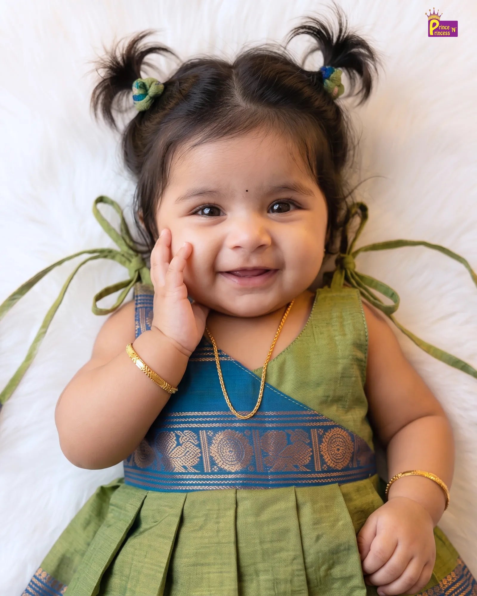 A smiling toddler girl wearing a traditional Indian dress, lying on a white faux fur surface. The dress is olive green with a blue and gold zari embroidered bodice.