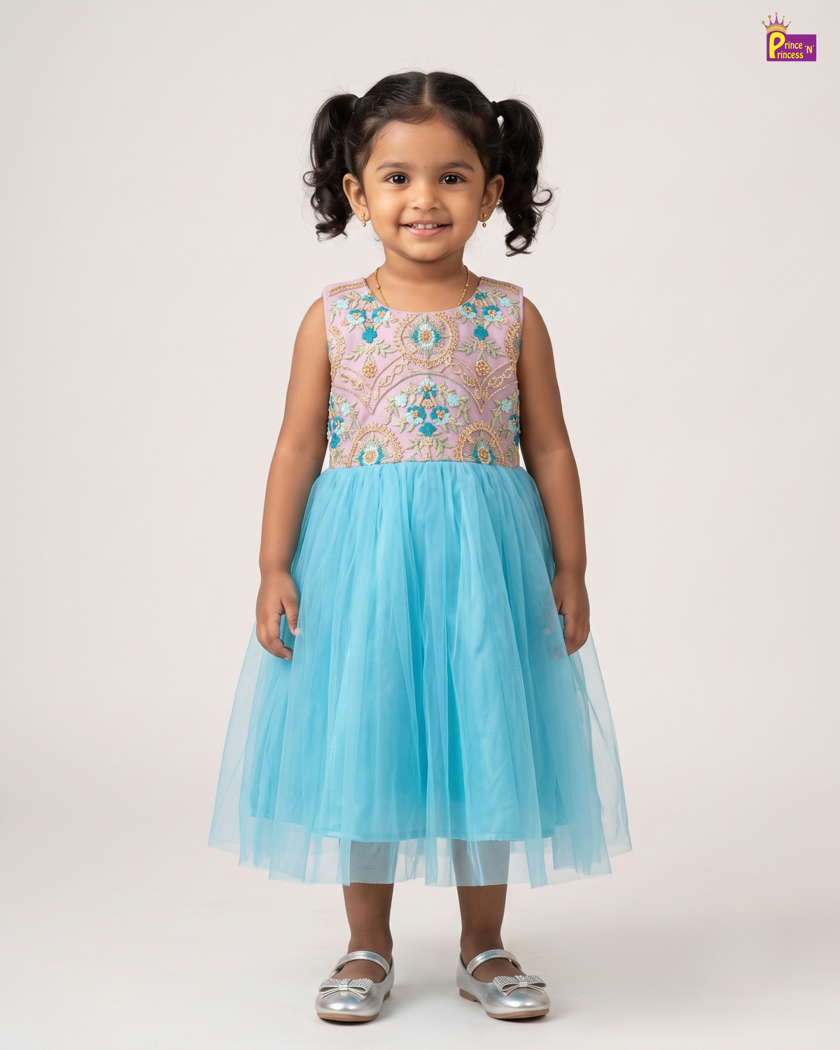 A young girl with dark curly hair in pigtails stands against a light beige studio background wearing a sleeveless tulle dress with a multicolored embroidered bodice and a light blue tulle skirt, paired with silver shoes.