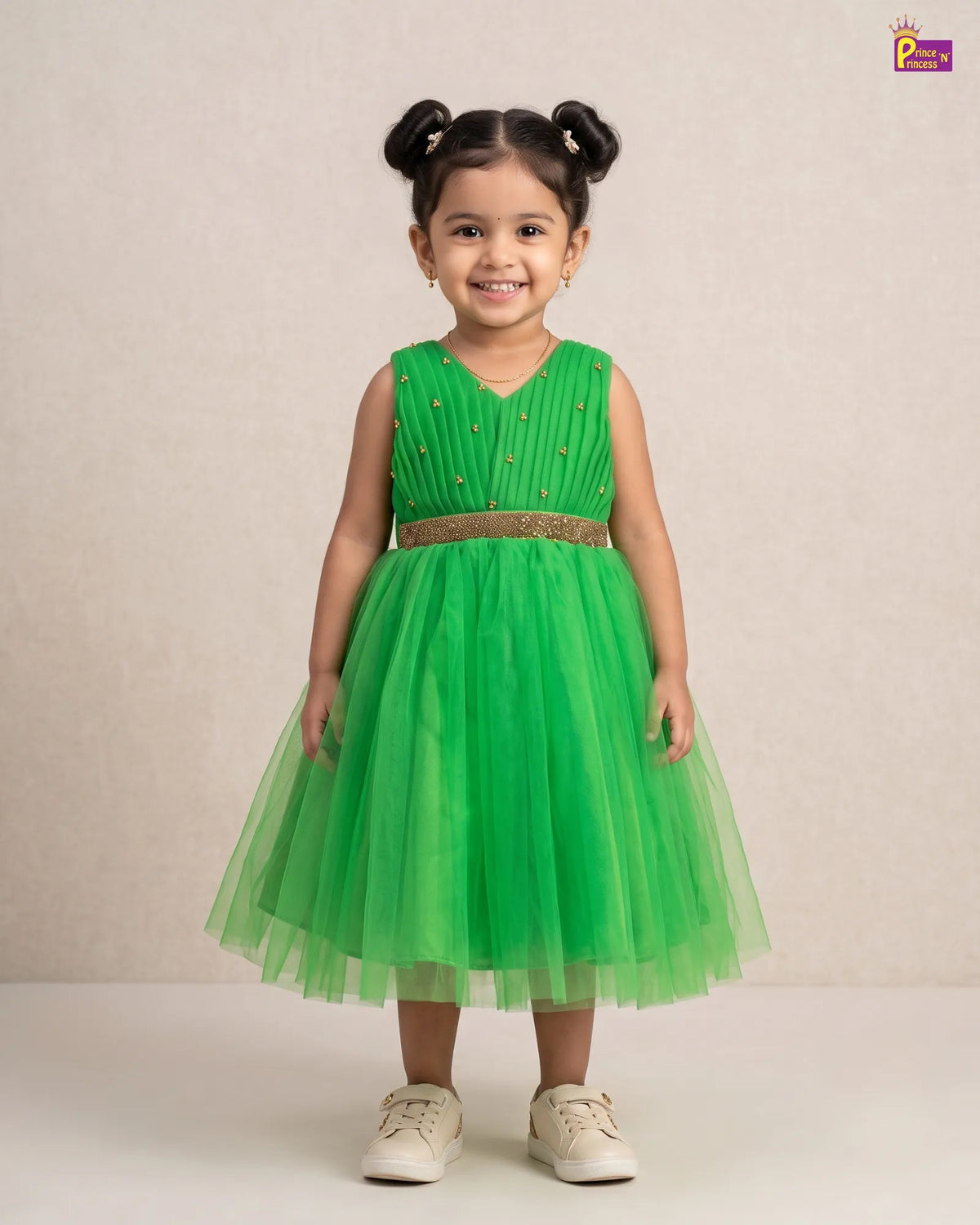 A young girl with her hair in two buns stands wearing a sleeveless bright green party dress with a pleated bodice, a glittery gold waistband, and a layered tulle skirt. She is wearing off-white sneakers against a neutral beige studio background.
