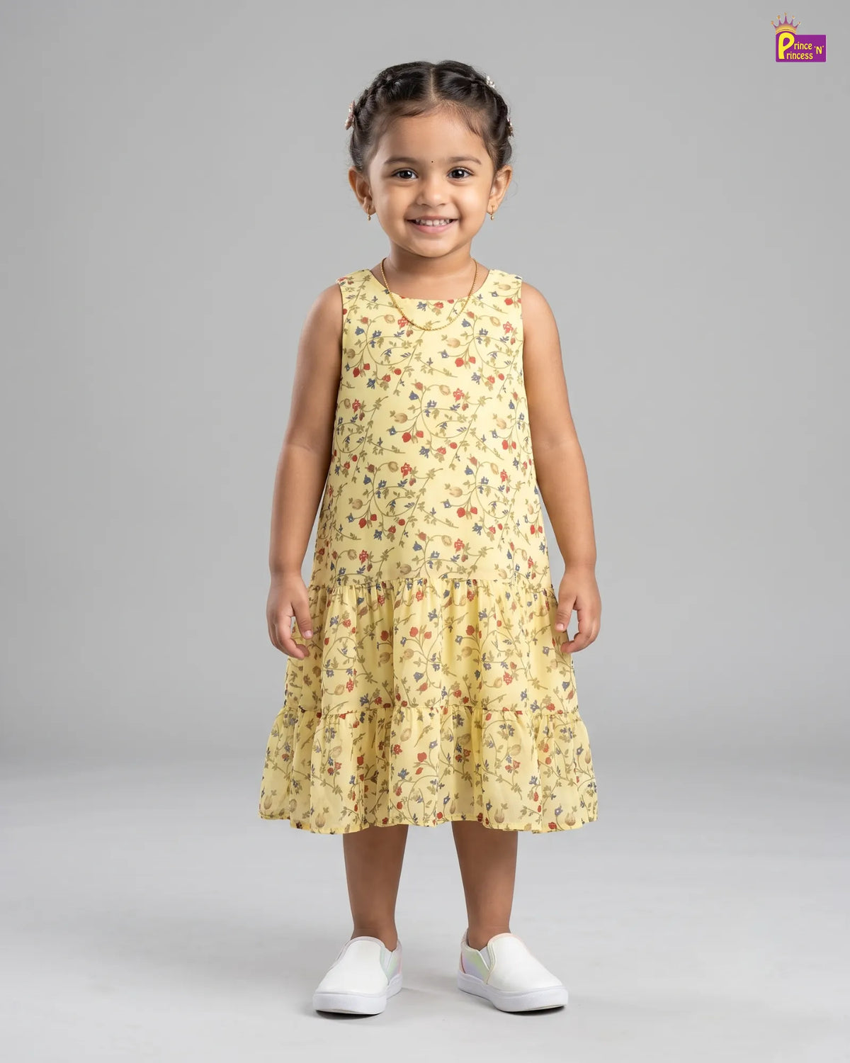 A young girl with braided hair stands against a light gray studio background wearing a sleeveless tiered yellow dress with a small multicolor floral print and white slip-on shoes.