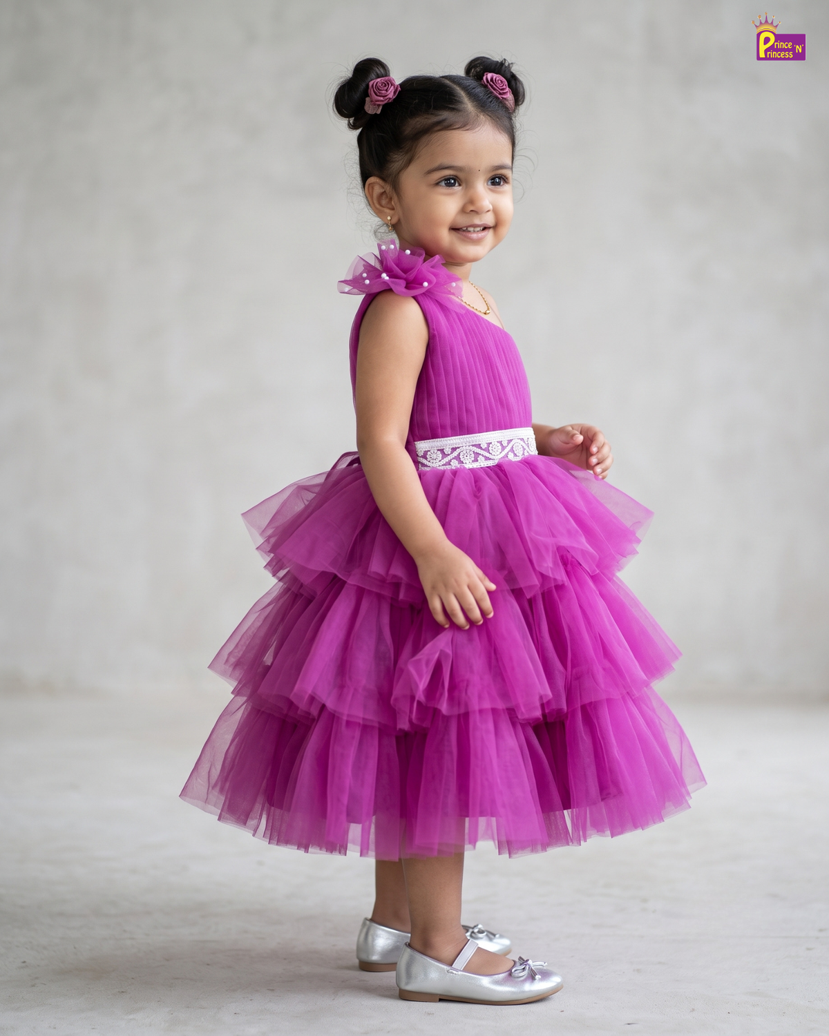 A young girl with dark hair in two buns wearing a bright magenta tiered tulle dress with a white embroidered waist sash, standing against a neutral gray studio background.