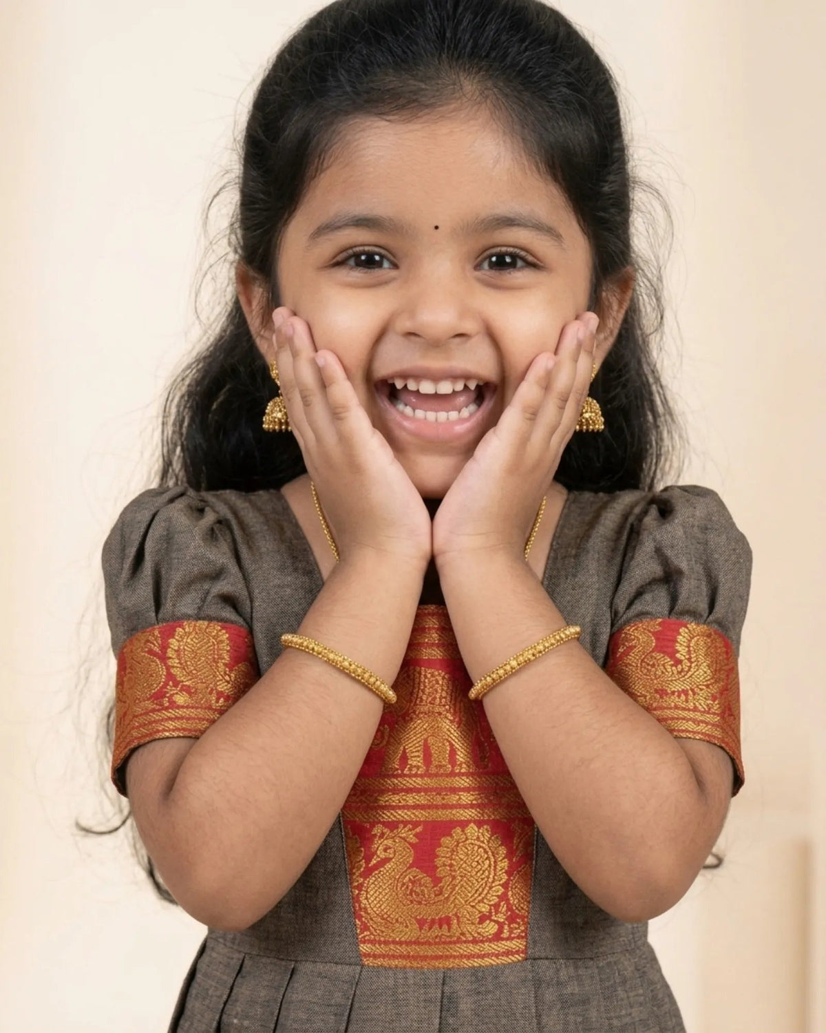 A young girl with dark hair smiling broadly, wearing a traditional Indian outfit with a brown body and red and gold embroidered borders, along with gold jewelry.