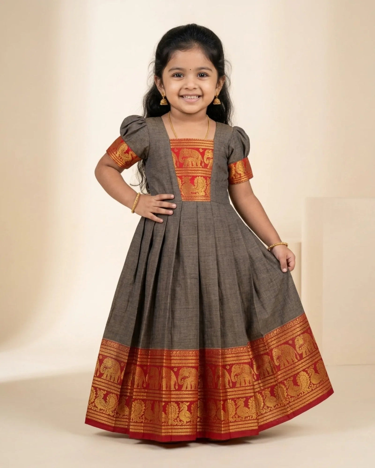 A young girl modeling a dark gray Anarkali-style dress with red and gold elephant-patterned borders and matching embroidered sleeves, set against a plain beige studio background.
