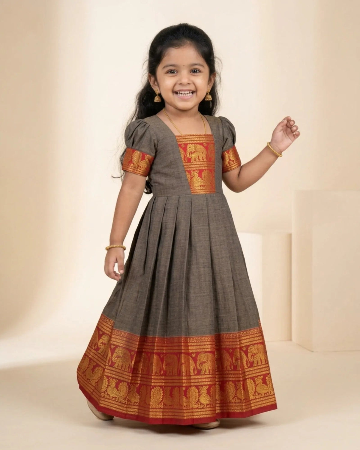 A young girl smiling and wearing a dark gray cotton A-line dress with red and gold elephant-patterned borders and sleeve details, set against a warm beige studio background.