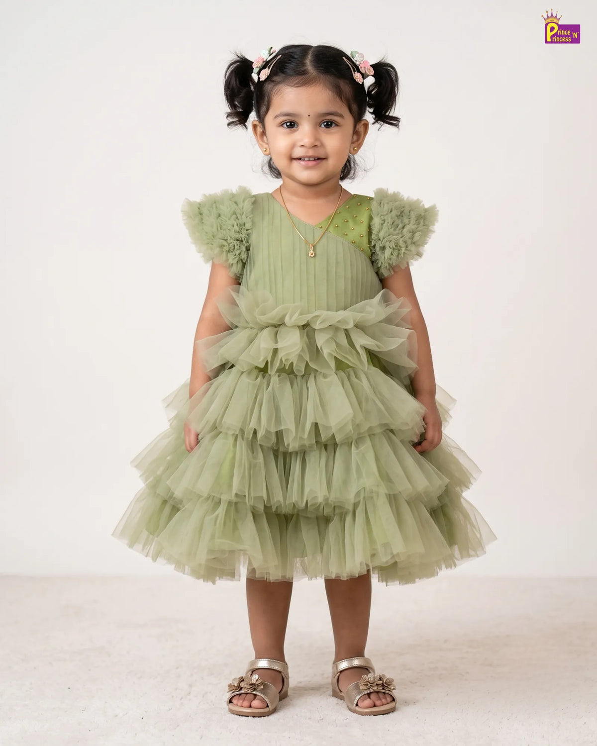 A young girl with dark hair in pigtails stands wearing a light sage green tiered tulle dress with ruffled shoulder details and metallic sandals, photographed against a plain off-white studio background.
