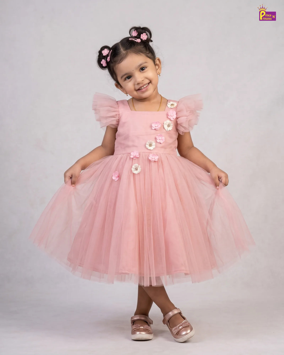 A young girl with dark hair styled in two buns wearing a pink tulle dress with 3D floral embellishments, standing against a light gray studio background.