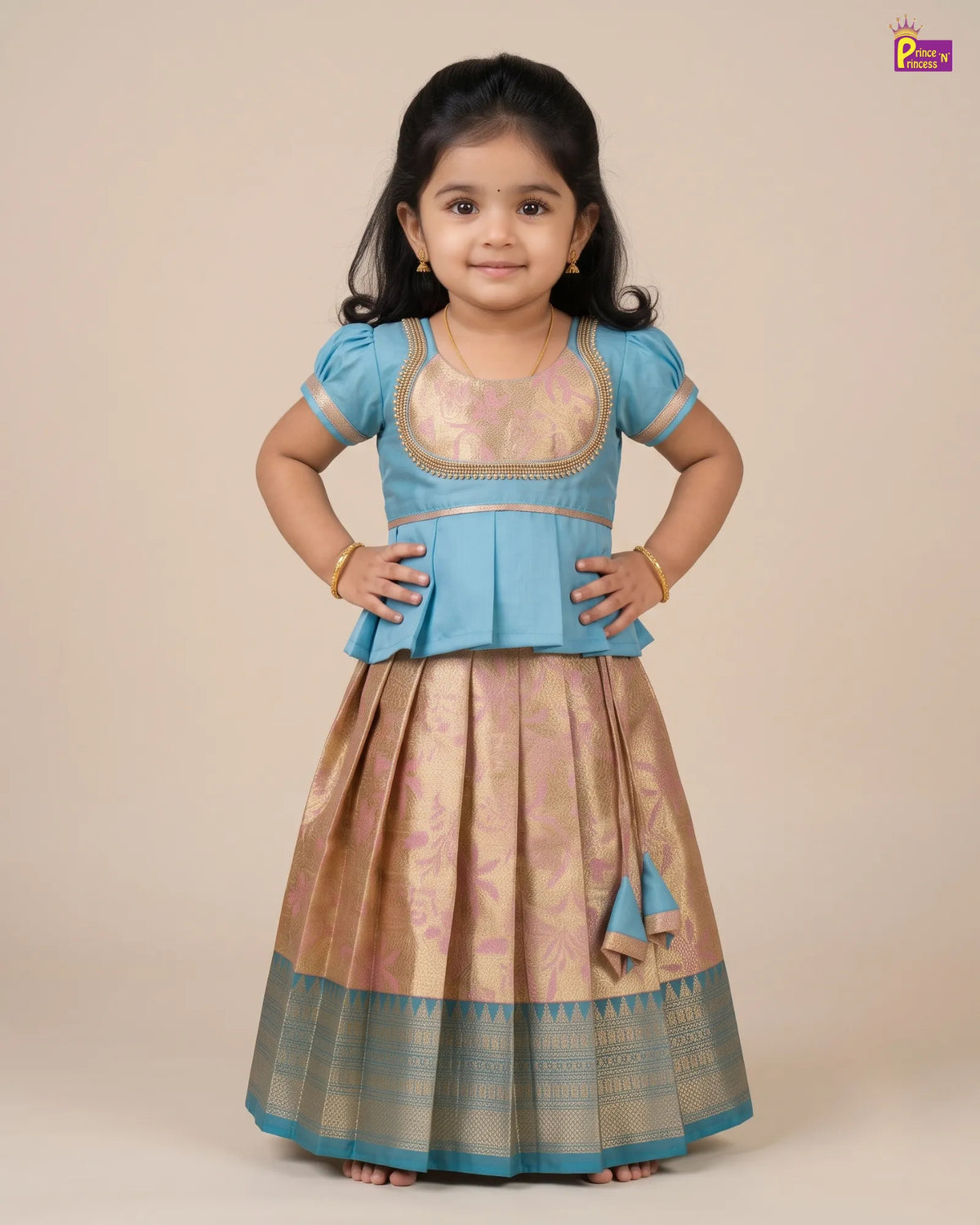 A young girl modeling a traditional Indian outfit consisting of a light blue short-sleeve blouse with gold embroidery and a matching pleated beige skirt with a blue border, set against a solid beige background.
