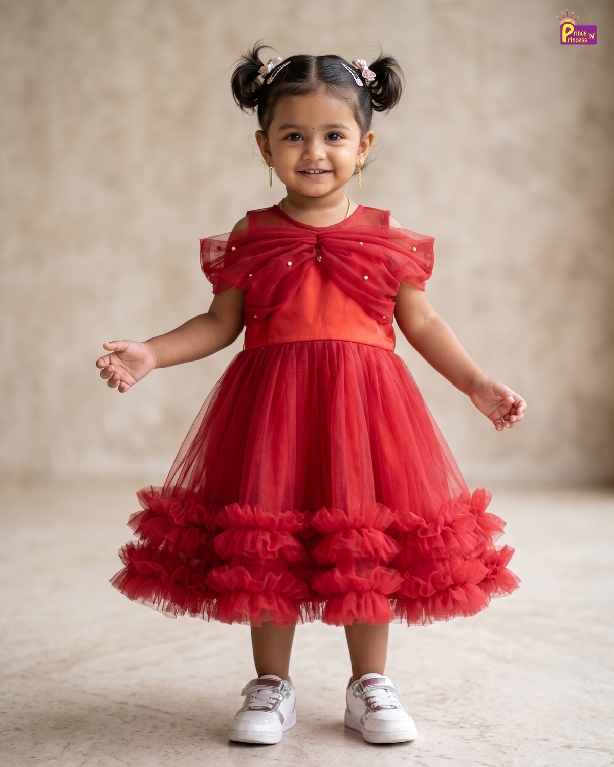 A toddler girl with her hair in two buns wearing a bright red party dress with a ruffled tulle skirt and white sneakers, standing against a neutral beige studio backdrop.
