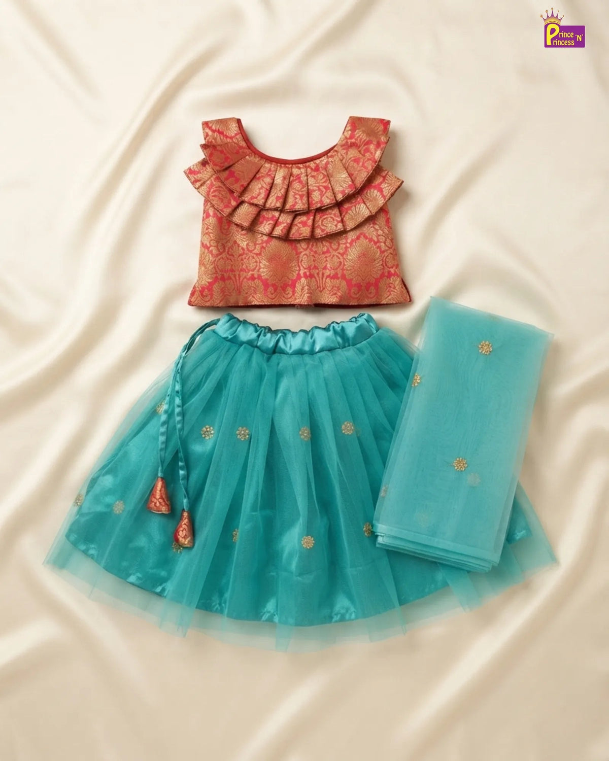 A children's traditional outfit set laid flat on a cream satin fabric background, featuring a coral-pink ruffled blouse with gold motifs, a turquoise blue tulle skirt with gold floral embroidery and a tassel tie, and a matching folded turquoise dupatta.