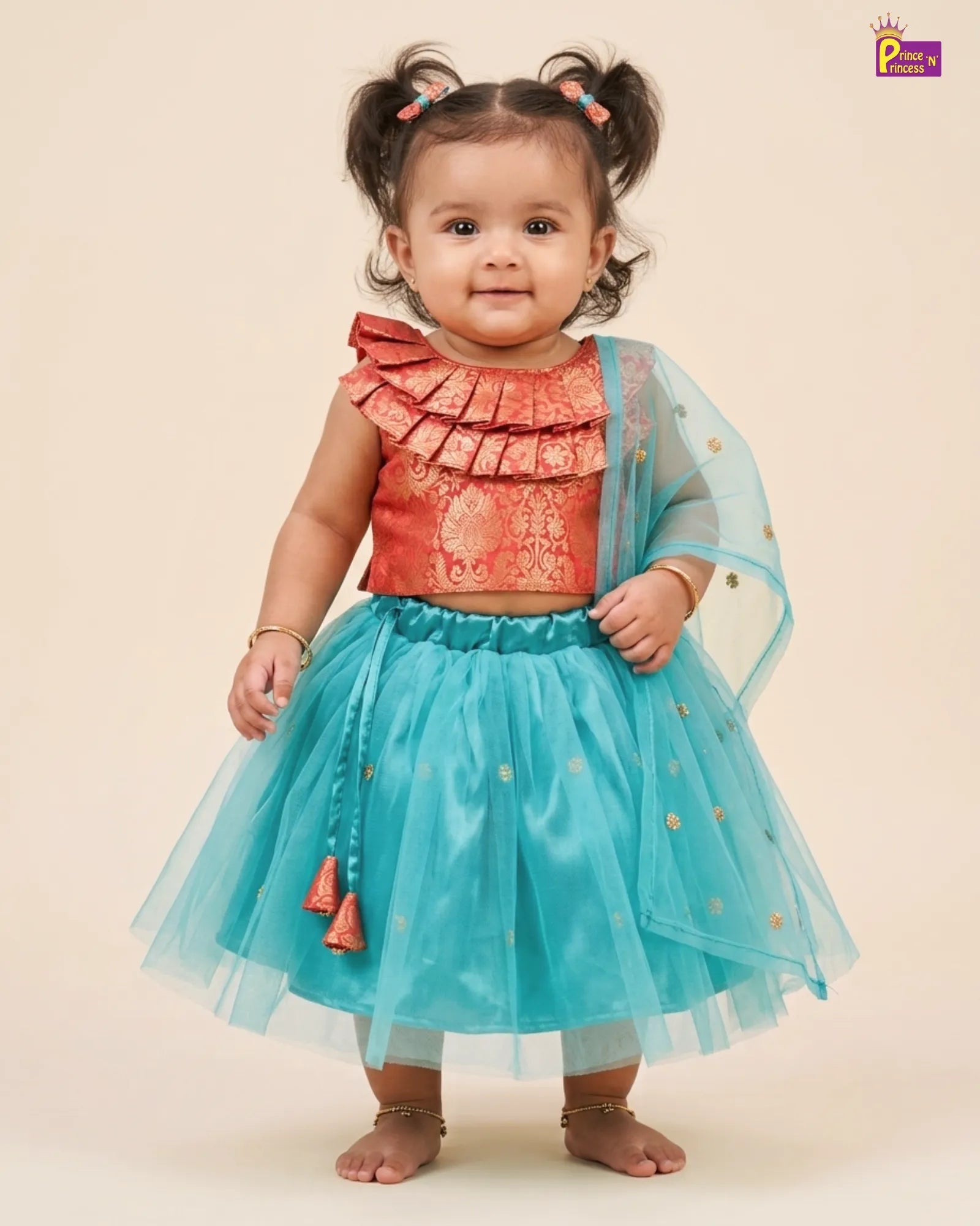A toddler girl wearing a traditional Indian ethnic outfit with a coral orange patterned blouse featuring ruffles and a turquoise blue tulle skirt with gold polka dots and a matching sheer dupatta, posed against a solid beige background.